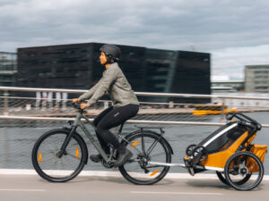 Cyclist wearing a helmet tows a Thule Chariot bike trailer along a waterfront path in a city.
