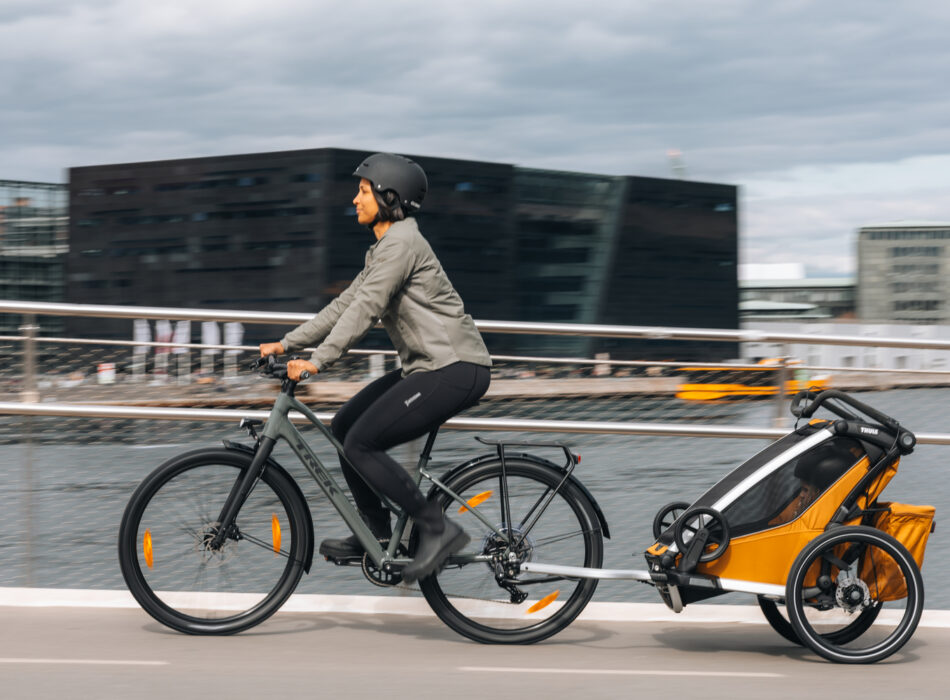 Cyclist wearing a helmet tows a Thule Chariot bike trailer along a waterfront path in a city.