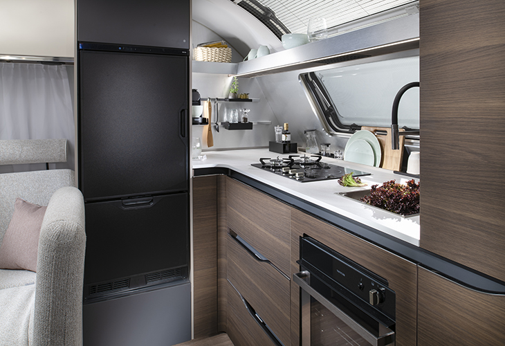 Adria Alpina 2027 caravan kitchen with black fridge-freezer, gas hob, oven and wood-finish cabinetry