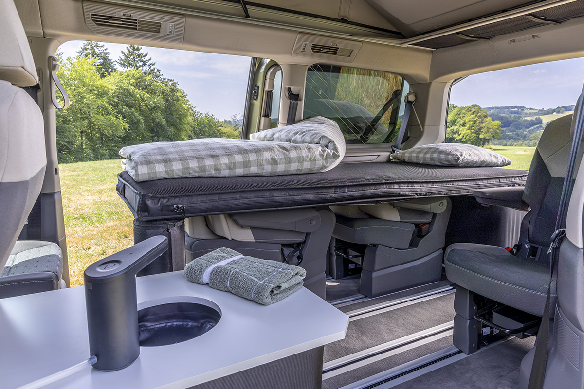 Volkswagen California camper interior with elevated bed, fold-down seats, and compact sink unit with towel