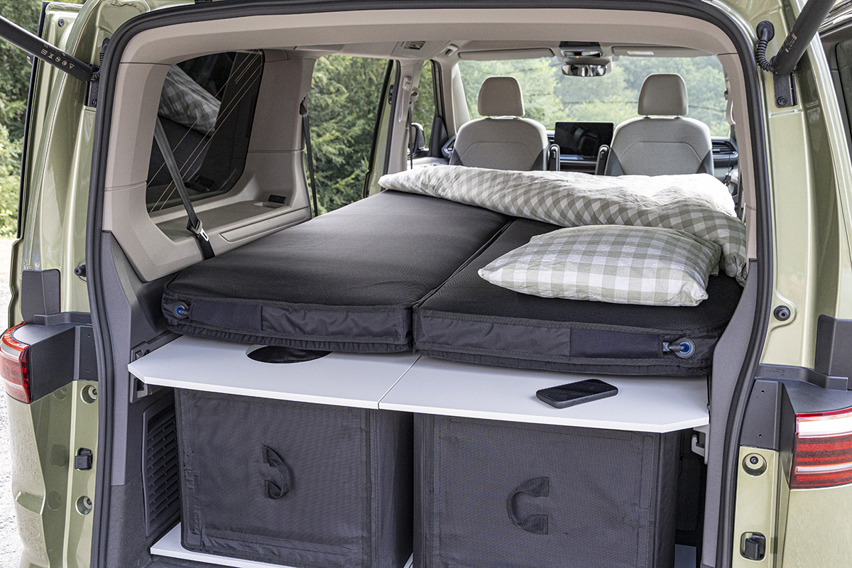 VW California camper van boot with folded bed, checked bedding, and storage boxes beneath a fold-out table.