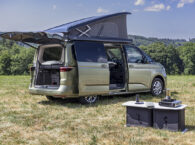 Volkswagen California camper van with elevating roof and open doors, alongside portable outdoor kitchen unit on grass field.