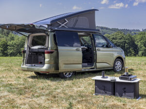 Volkswagen California camper van with elevating roof and open doors, alongside portable outdoor kitchen unit on grass field.