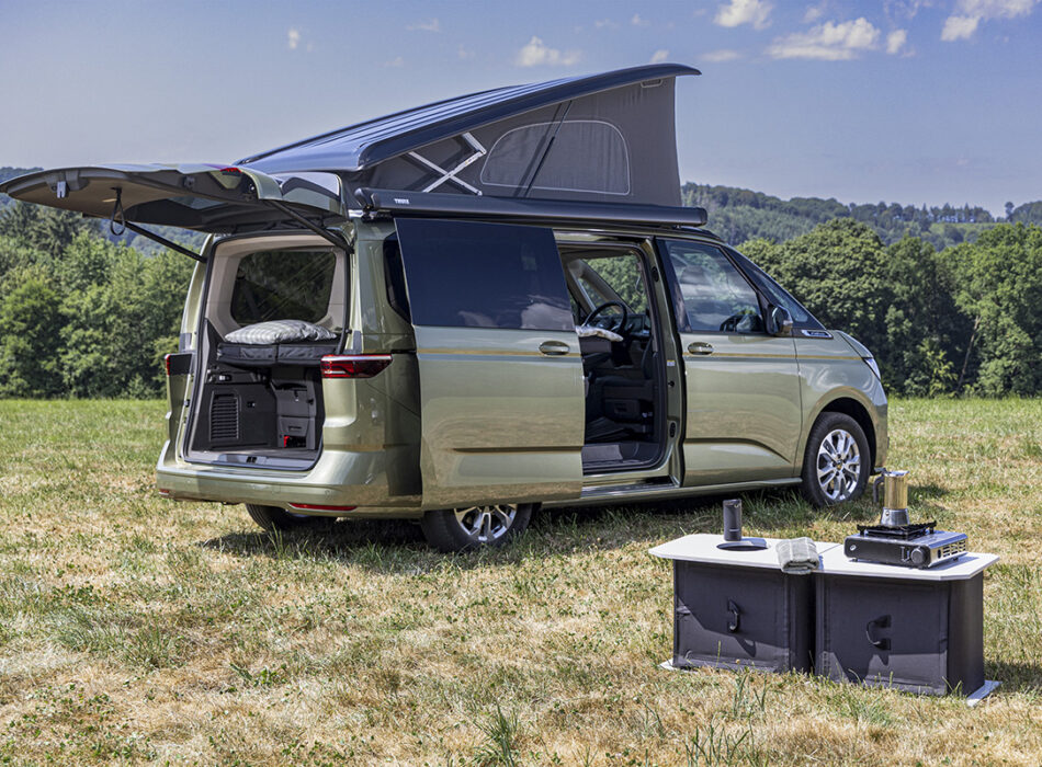 Volkswagen California camper van with elevating roof and open doors, alongside portable outdoor kitchen unit on grass field.