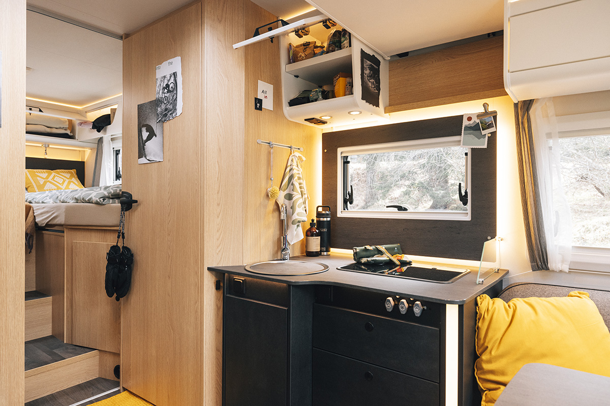 Compact kitchen in Sunlight UNLTD motorhome with sink, hob, storage cupboards and cosy sleeping area behind.