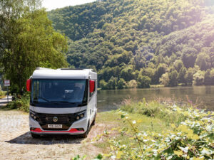 Niesmann+Bischoff iSmove 2027 motorhome parked beside a wooded riverbank, showcasing its sleek white and red exterior.