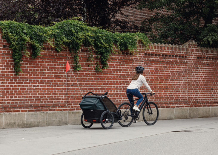 Cyclist riding beside a brick wall, towing a Thule Chariot bike trailer with an orange safety flag.