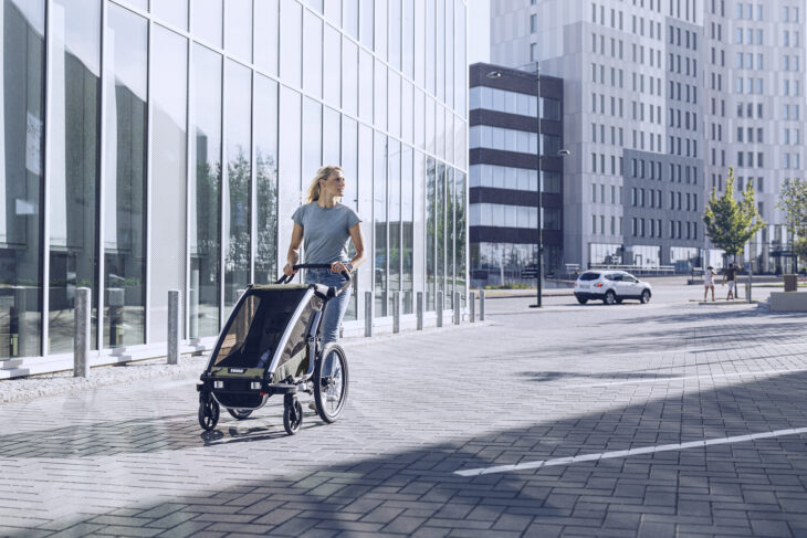 Woman walking with Thule Chariot bike trailer on paved urban plaza beside glass office buildings