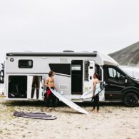 Two surfers unload boards from a Sunlight UNLTD motorhome parked on a beach with cliffs behind.