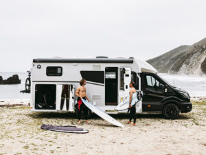 Two surfers unload boards from a Sunlight UNLTD motorhome parked on a beach with cliffs behind.