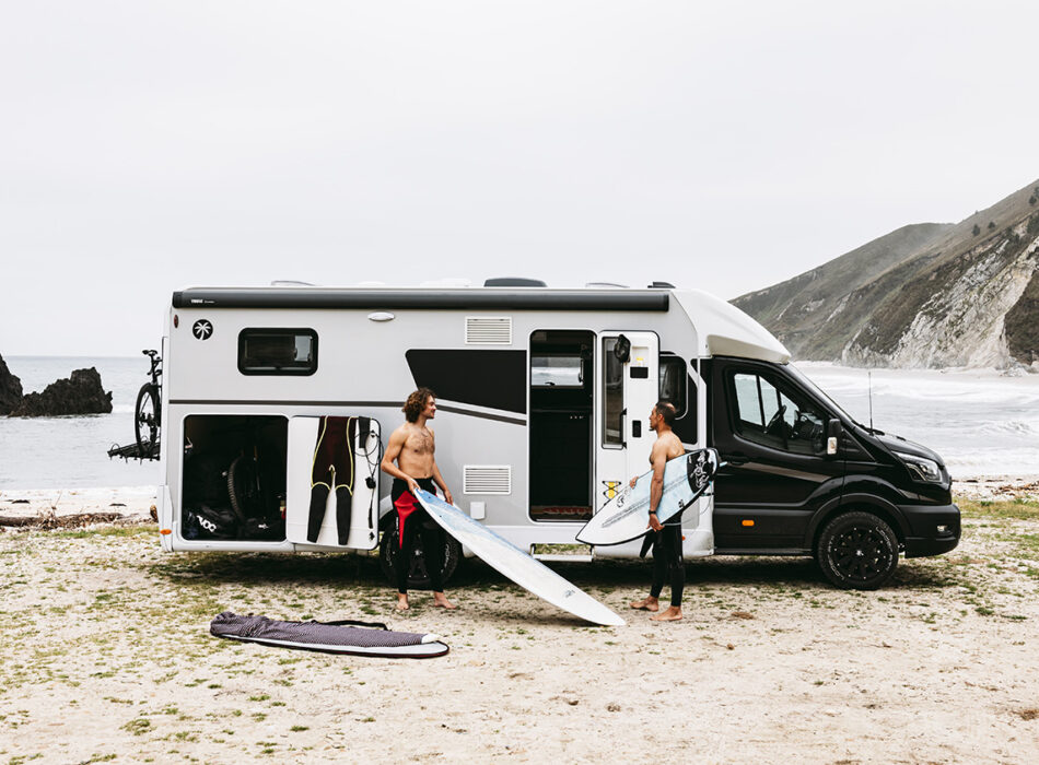 Two surfers unload boards from a Sunlight UNLTD motorhome parked on a beach with cliffs behind.
