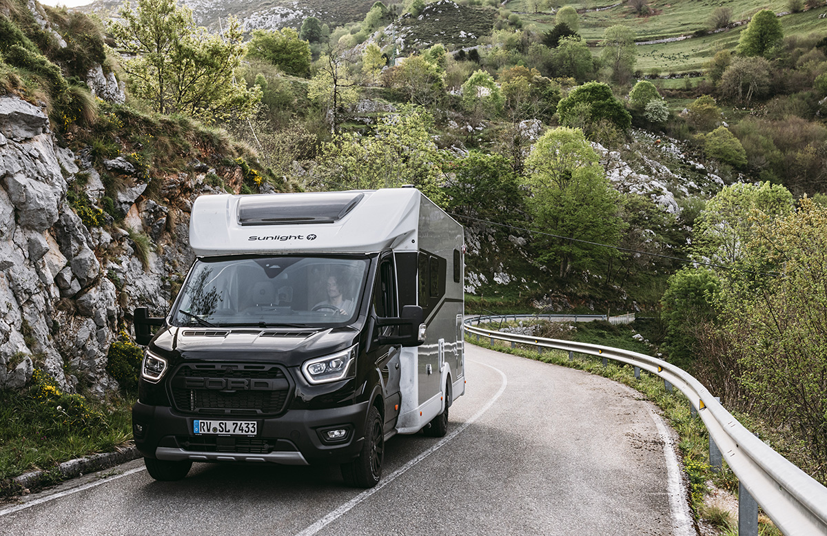 Sunlight UNLTD motorhome on Ford chassis navigating a winding mountain road through rocky, green terrain.