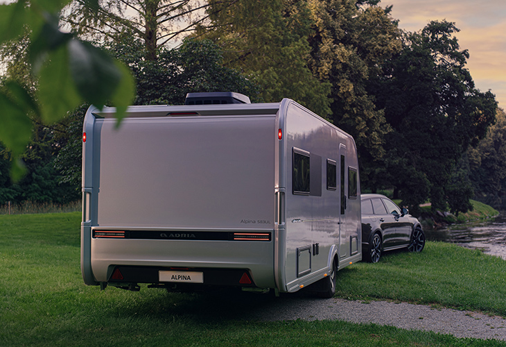 Adria Alpina 2027 caravan rear view, towed by dark SUV, parked on grass beside riverside trees at dusk
