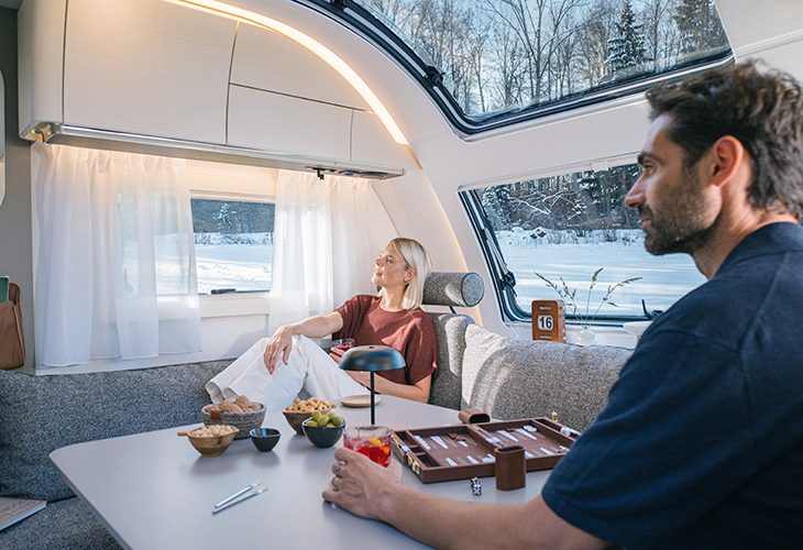 Adria Alpina 2027 winter dinette interior with couple relaxing, backgammon board and snowy view through window