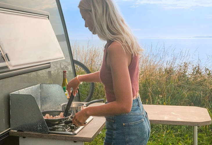 Woman cooking on the Kip Kompakt caravan's outdoor kitchen stove, with sea view in background.
