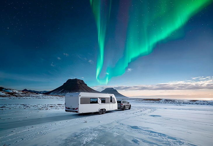 Knaus Nordwind caravan towed by SUV on snowy terrain under the northern lights, with mountains in the background.
