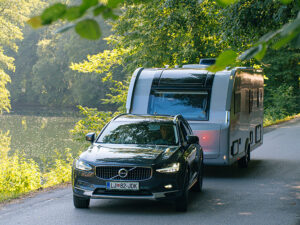 Volvo SUV towing the redesigned Adria Alpina 2027 caravan along a tree-lined road beside a lake