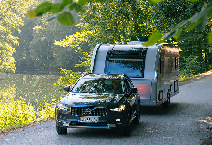 Volvo SUV towing the redesigned Adria Alpina 2027 caravan along a tree-lined road beside a lake