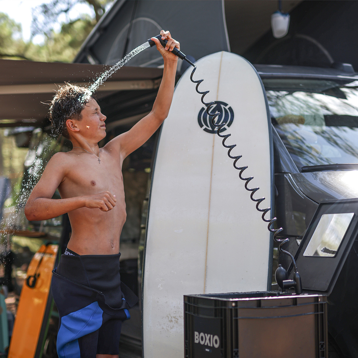 Boy rinsing off with portable shower head connected to Boxio XL washbasin unit beside surfboard and camper van