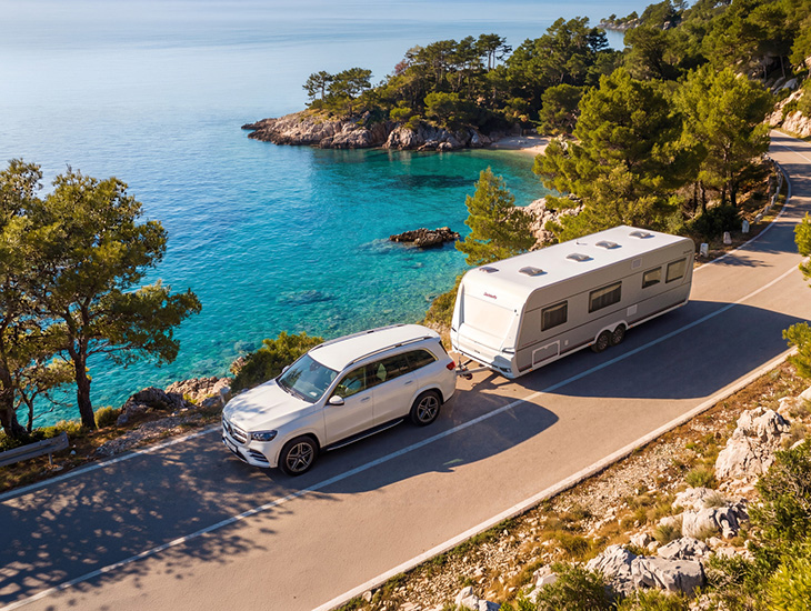 White SUV towing the Dethleffs Summer Edition 730 FKR caravan along a coastal road above turquoise sea.