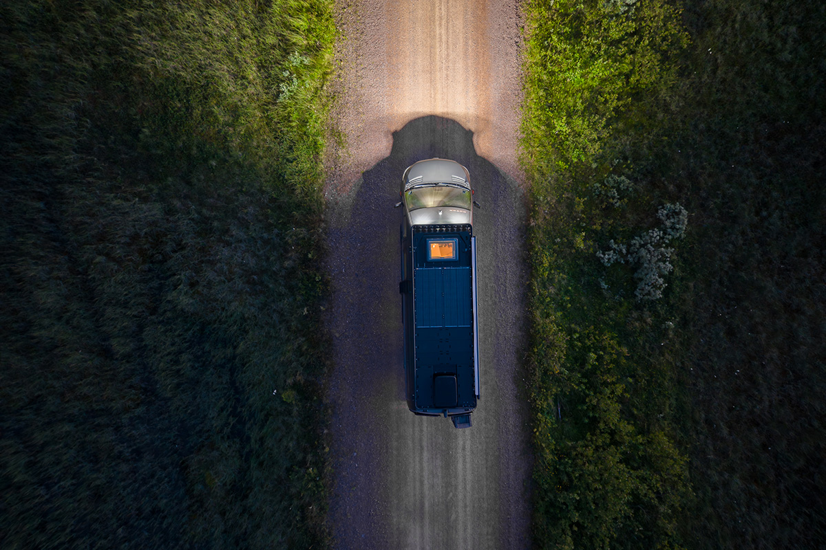 Aerial view of Dovra RIG X-710 camper van with roof solar panels driving along a tree-lined gravel road at dusk.