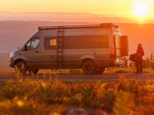 Dovra RIG X-710 camper van parked at sunset, rear doors open, man unloading gear from storage compartment