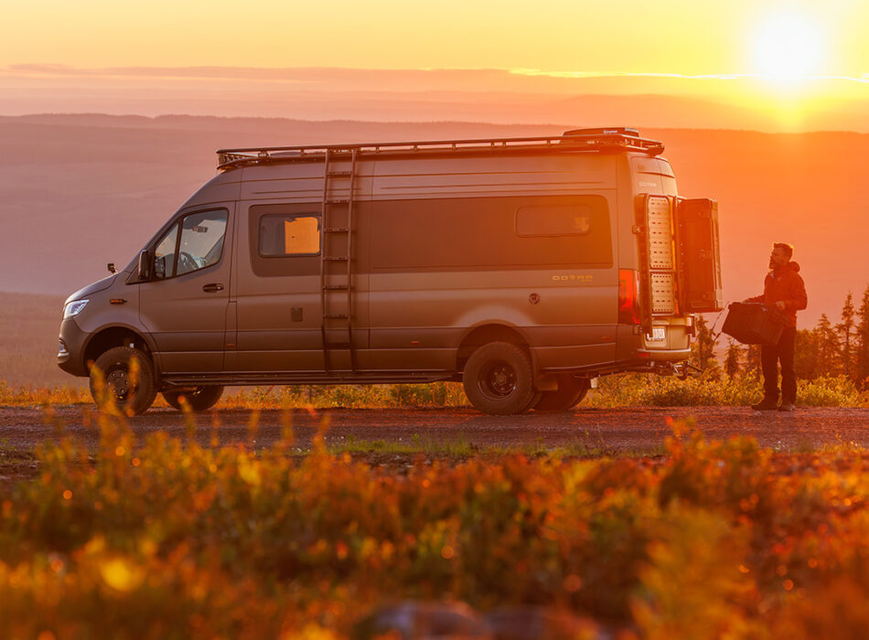 Dovra RIG X-710 camper van parked at sunset, rear doors open, man unloading gear from storage compartment