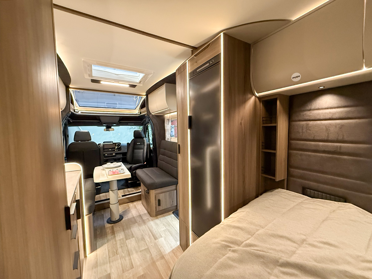 Kabe Novum Compact 720 LXL motorhome interior showing bed, driving cab, dinette and wood-finish cabinetry