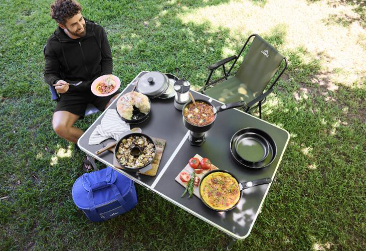 Man eating camping breakfast at folding table with Lidl camping deals: pans, chair, coffee pot and cool bag on grass